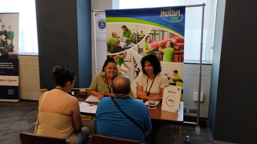 Dos personas sentadas frente a un escritorio, delante de una pancarta que dice 'Motiva CEE', atendiendo a dos personas sentadas de espaldas a la cámara frente al escritorio